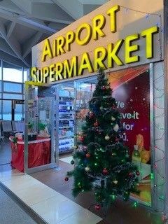 Beleuchtetes Schild 'AIRPORT SUPERMARKET' über einem Eingang mit Weihnachtsbaum im Vordergrund in einer Flughafenhalle