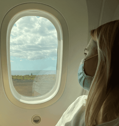 Frau mit Gesichtsmaske blickt aus dem Flugzeugfenster auf Wolken und Landschaft.