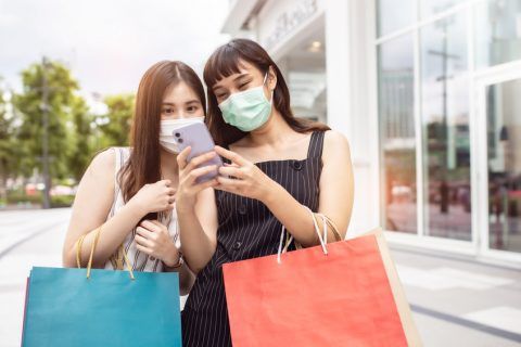 Zwei Frauen mit Einkaufstaschen und Gesichtsmasken betrachten gemeinsam ein Smartphone vor einem Geschäft